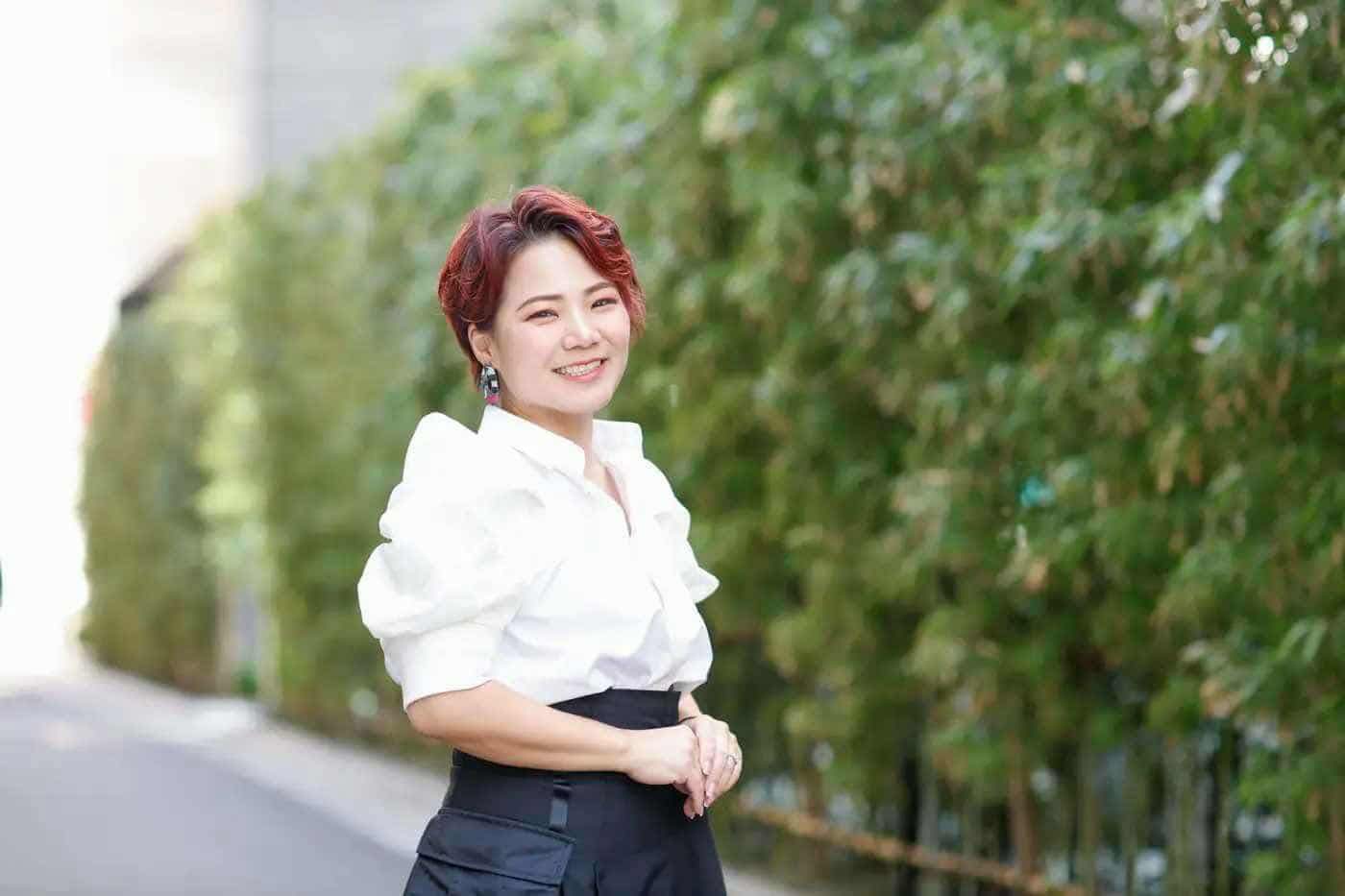 **Alt Text:** A woman with short, red hair, dressed in a white blouse and black pants, posing for a photograph in front of...